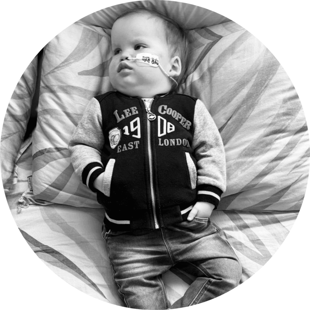 Ananda lies on pillows with infant jeans & a light jacket on. He's looking up at his family.
