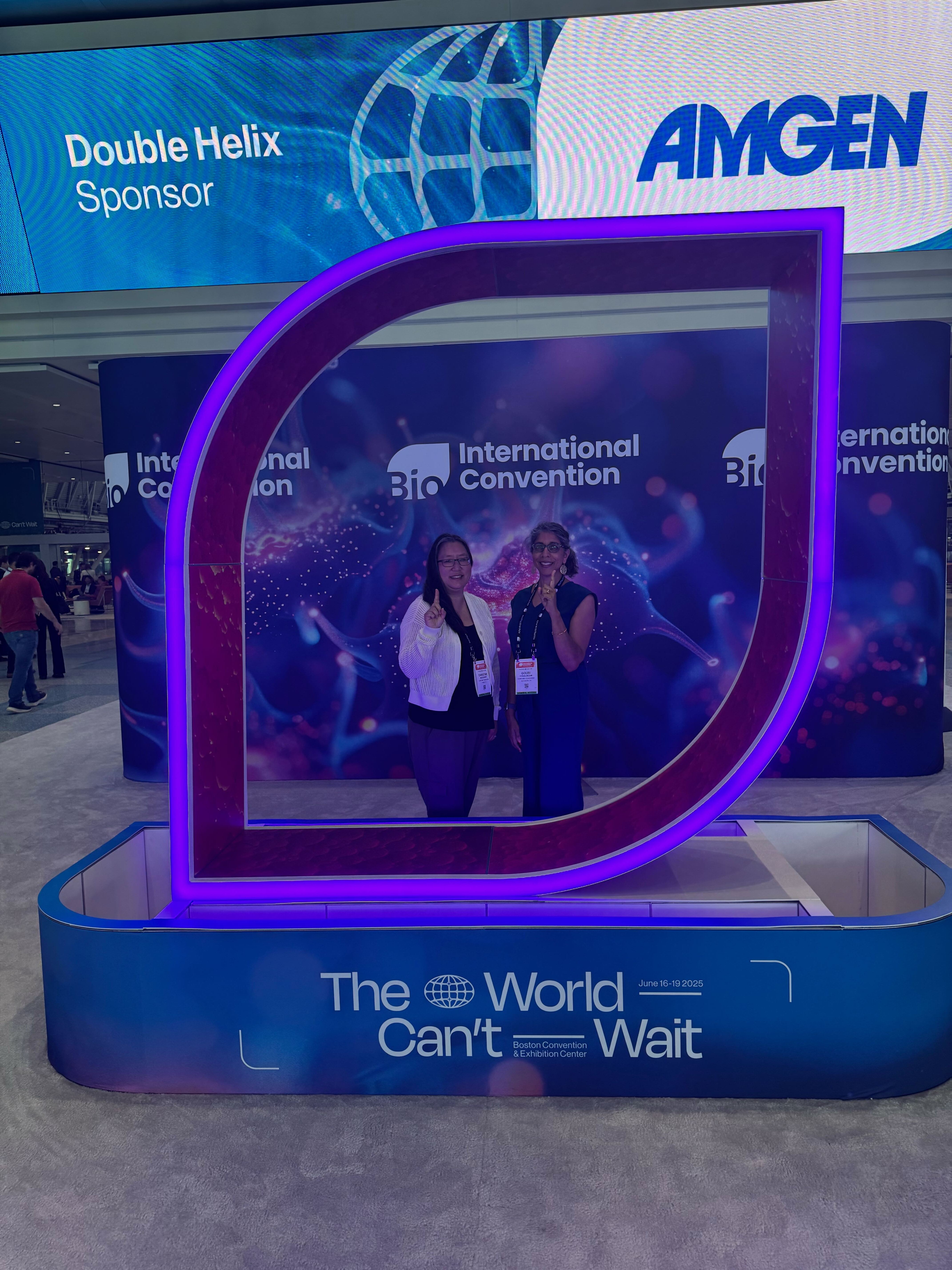 Christine and Gouri are shown at the BIO International conference in front of the Amgen exhibit.